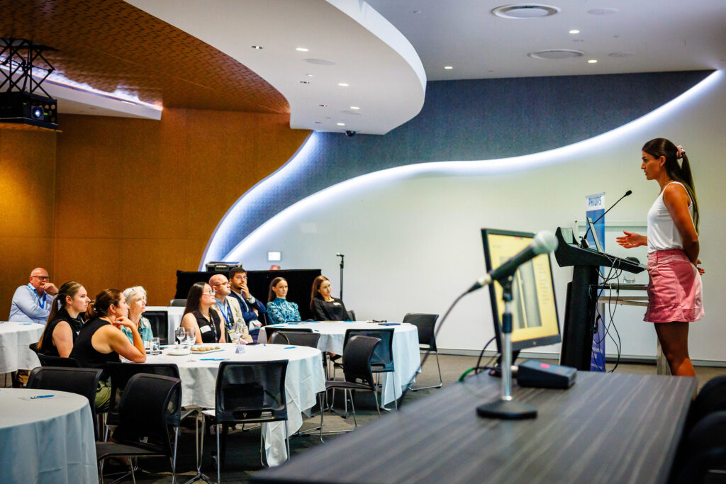 A woman is speaking at a podium to a crowd seated at round tables.
