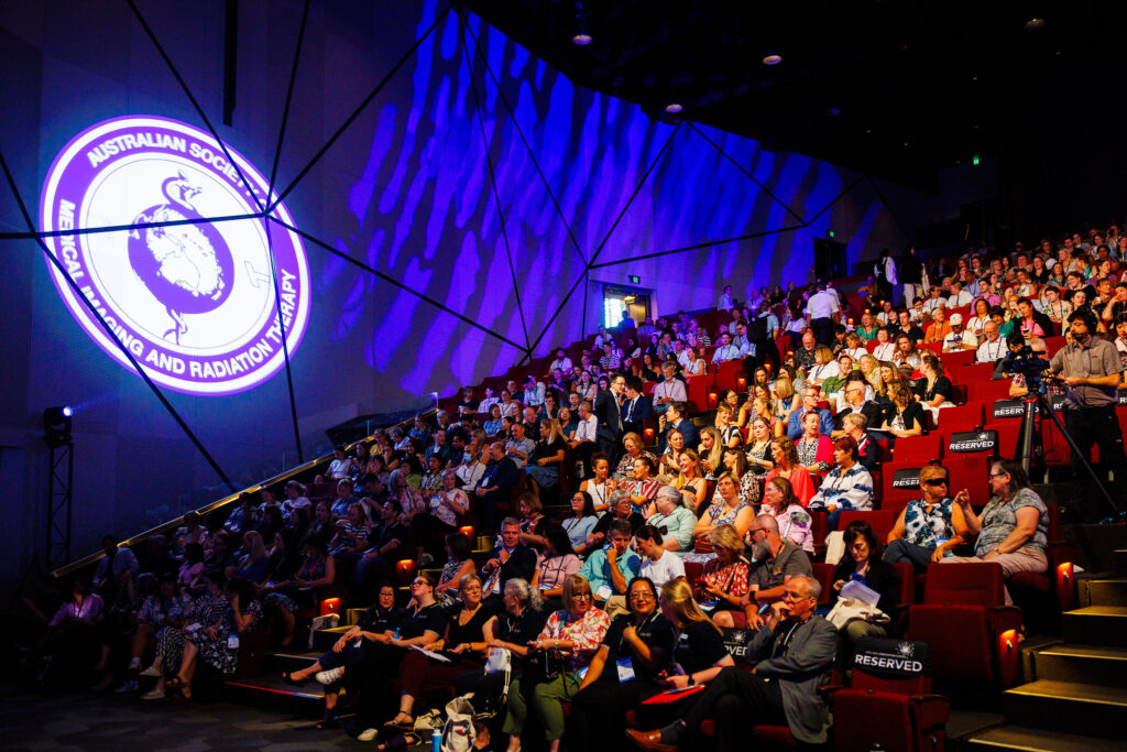 A large crown in an Auditorium. The ASMIRT logo is projected on the wall beside them.